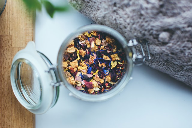 An open glass jar filled with a colorful mixture of dried flowers and herbs, placed on a white surface next to a grey fabric.