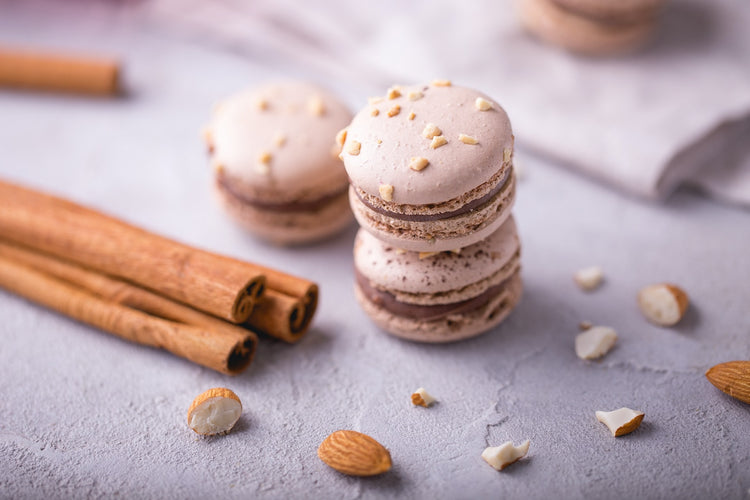 Three beige macarons with nuts on top are stacked on a light gray surface, surrounded by cinnamon sticks and scattered almond pieces.