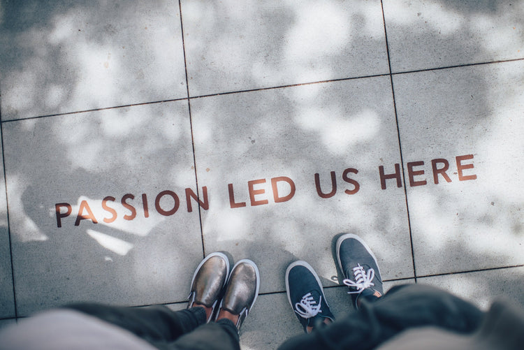 Two pairs of feet in casual shoes stand on a sidewalk with the words "PASSION LED US HERE" written on it.