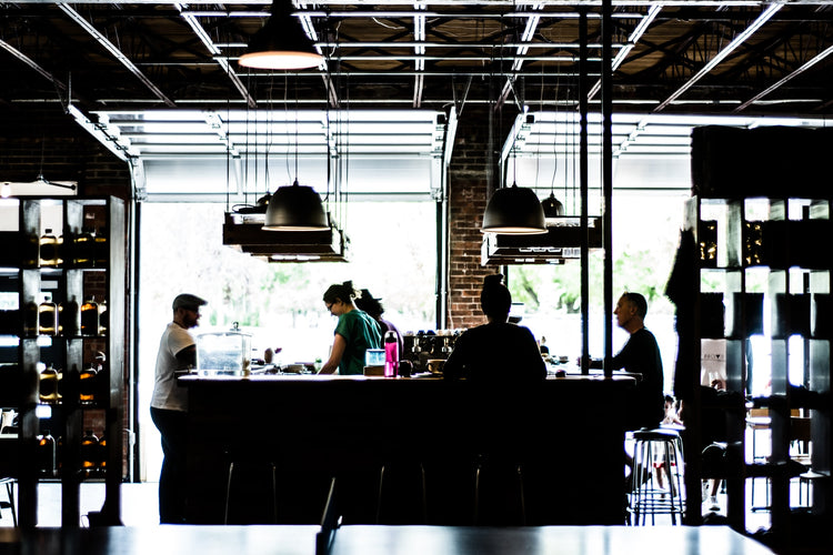 People are standing and sitting around a large counter inside a well-lit, modern cafe with a high ceiling and industrial-style lighting.