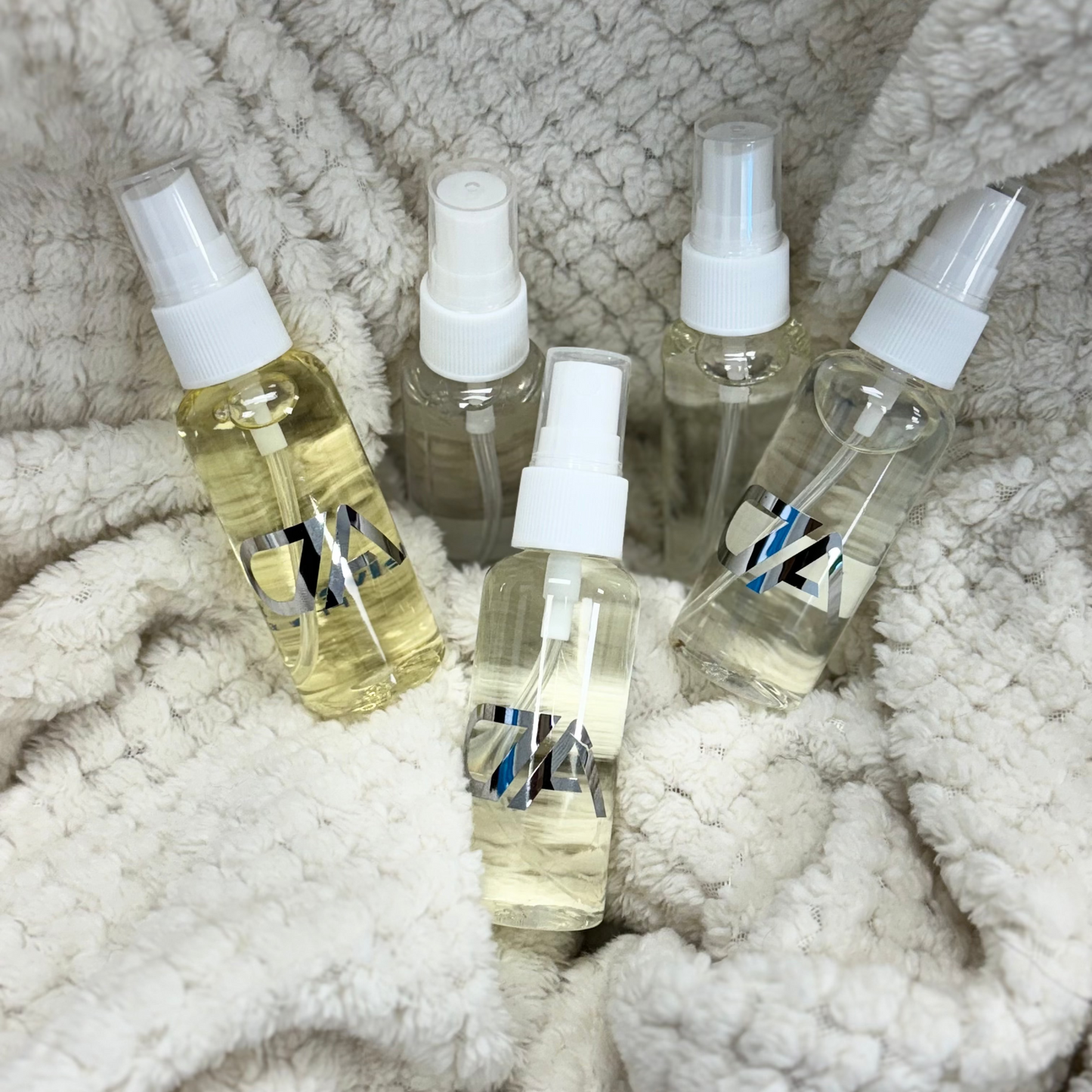 Five Mini Room Spray bottles with clear or light yellow liquid sit on a textured, cream-colored fabric, showcasing their compact, travel-friendly design for portable fragrance on the go.