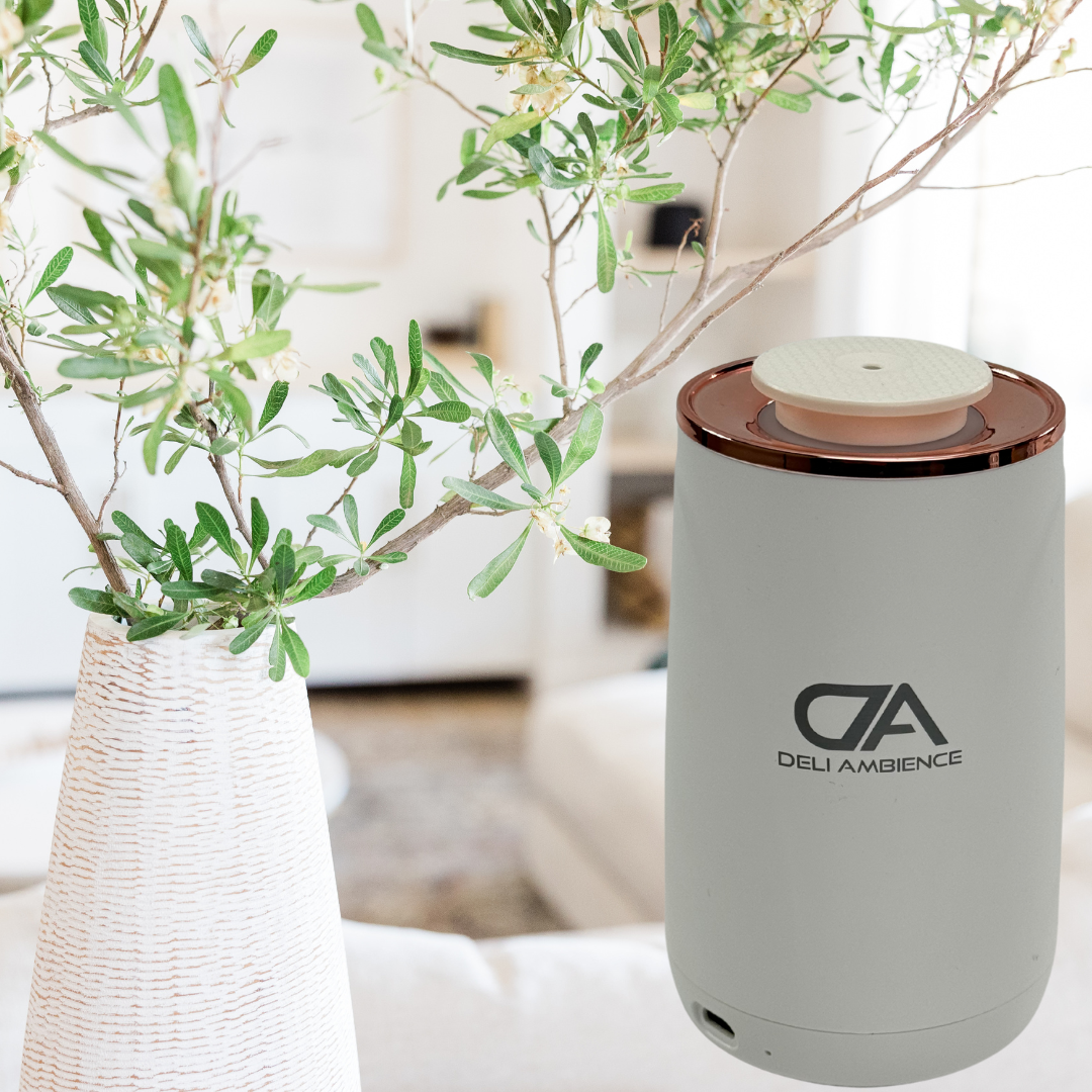 A grey air purifier by Deli Ambience is placed on a table next to a vase with green branches and the Venus Oil Diffuser with Sea Water Scent 100ml Bundle in a well-lit living room, creating the perfect setting for aromatherapy.