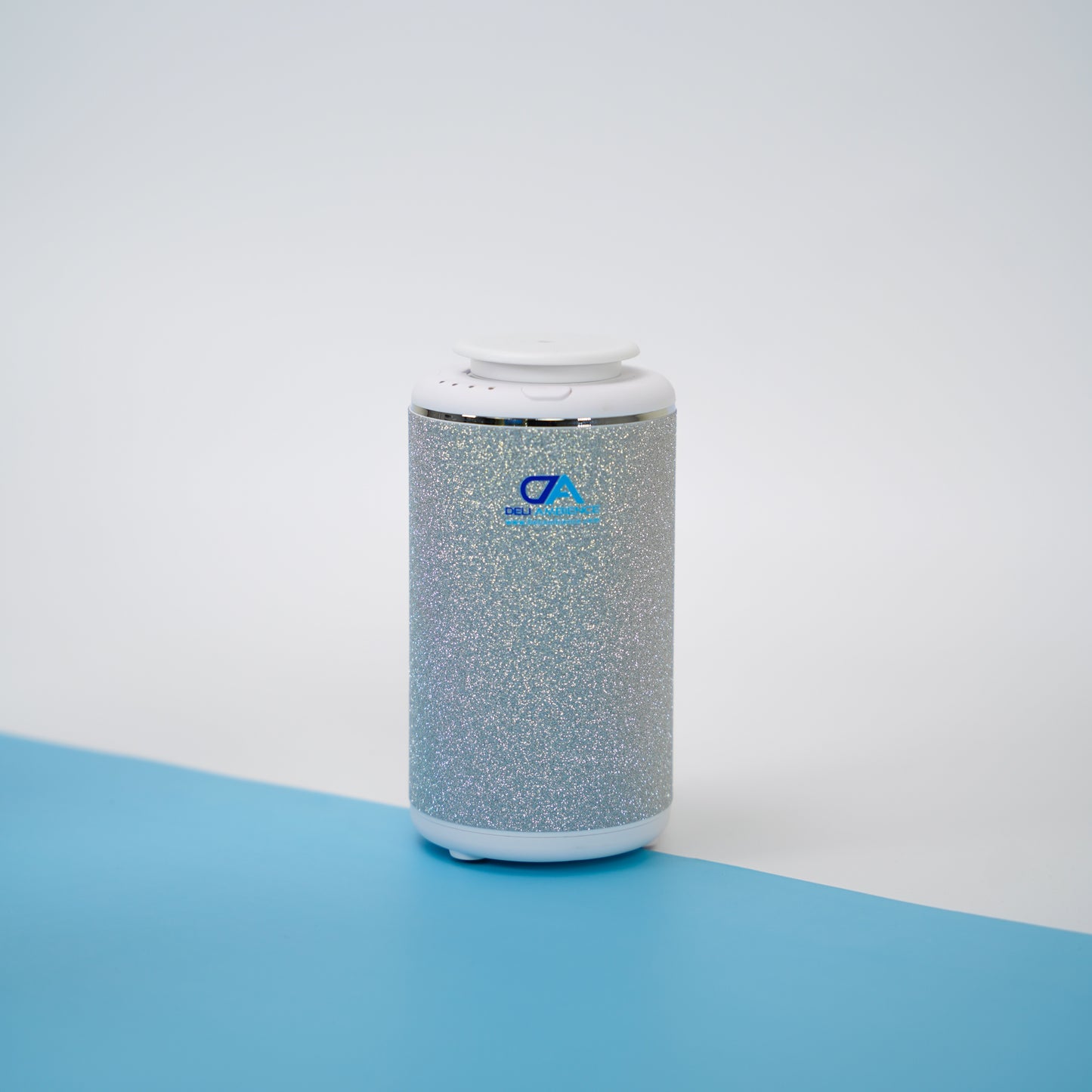 A Venus Compact Oil Diffuser USB-C Power, featuring a metallic silver cylindrical design with white accents on the top and bottom, rests on a blue and white surface, making it an ideal portable diffuser for small rooms.