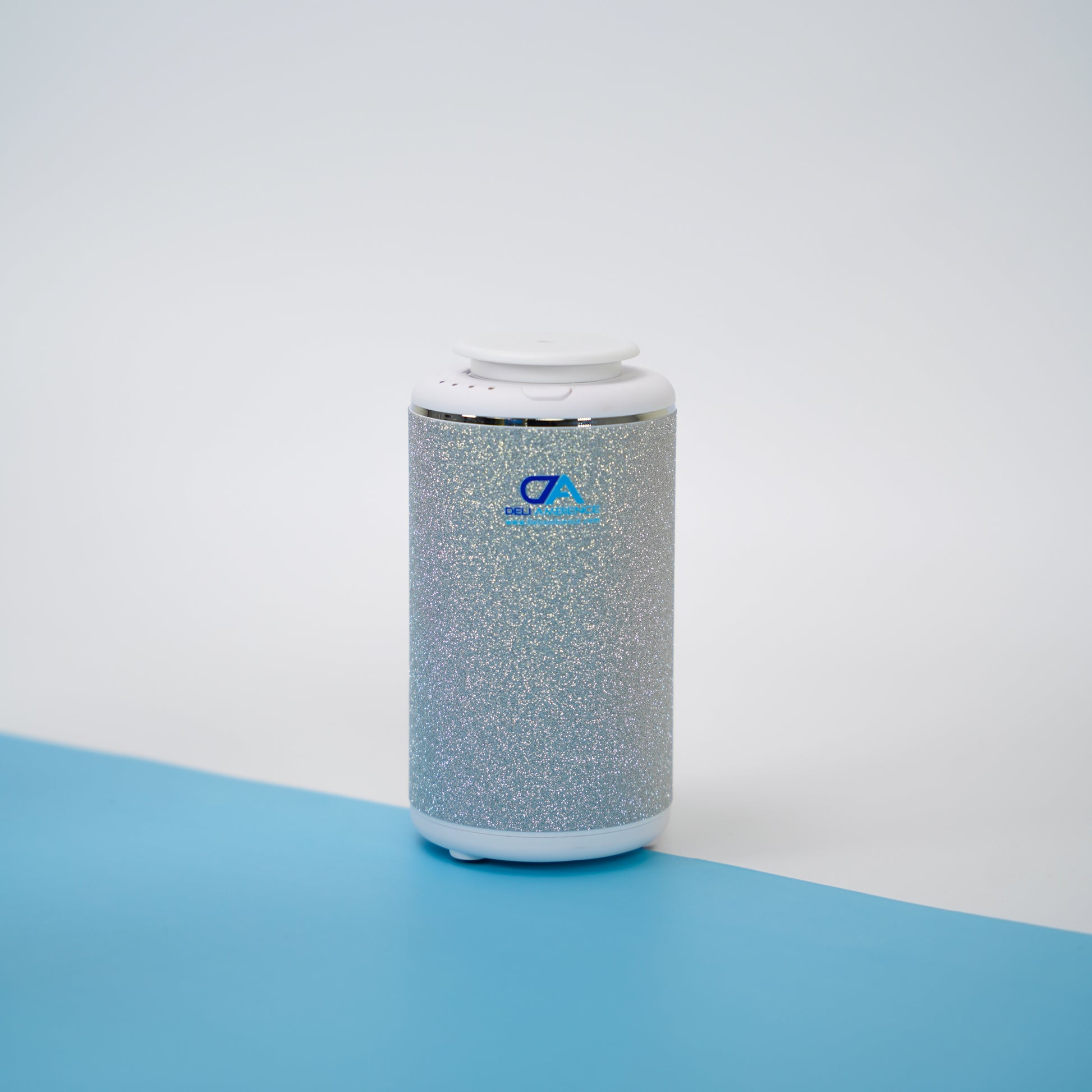 A Venus Compact Oil Diffuser USB-C Power, featuring a metallic silver cylindrical design with white accents on the top and bottom, rests on a blue and white surface, making it an ideal portable diffuser for small rooms.