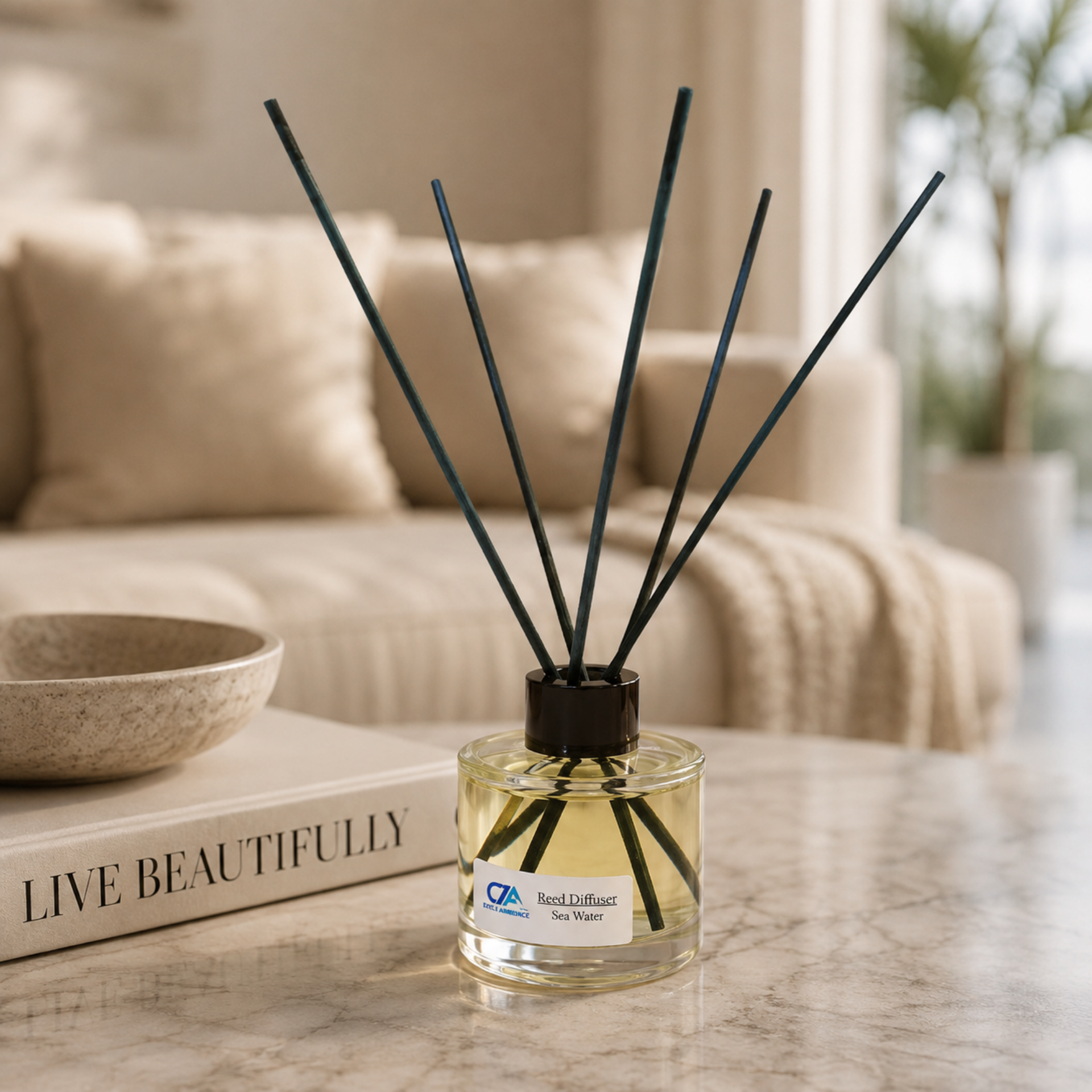 A bottle of Reed Diffusers filled with yellow liquid rests on a marble table beside a “Live Beautifully” book, subtly infusing the cozy living room with fragrance oils. A sofa and beige throw in the background enhance the inviting atmosphere, showcasing how Reed Diffusers serve as elegant natural air purifiers in home décor.