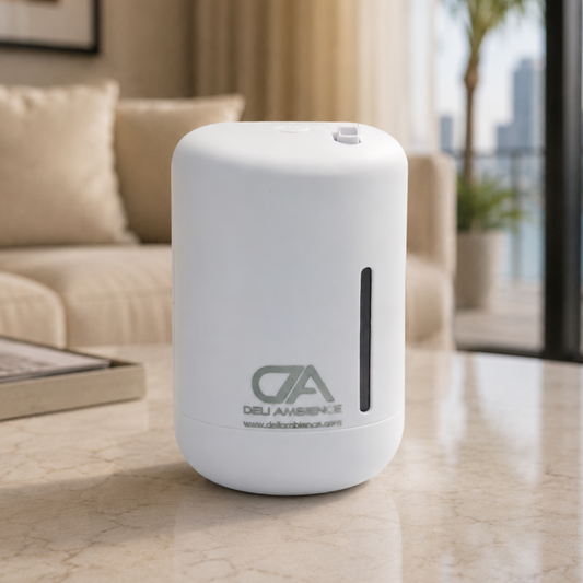An Afrodita Scent Diffuser, featuring a white cylindrical design and the “DELI AMBIENCE” logo, sits on a marble table in a living room. A sofa, a plant, and city buildings are visible in the background.